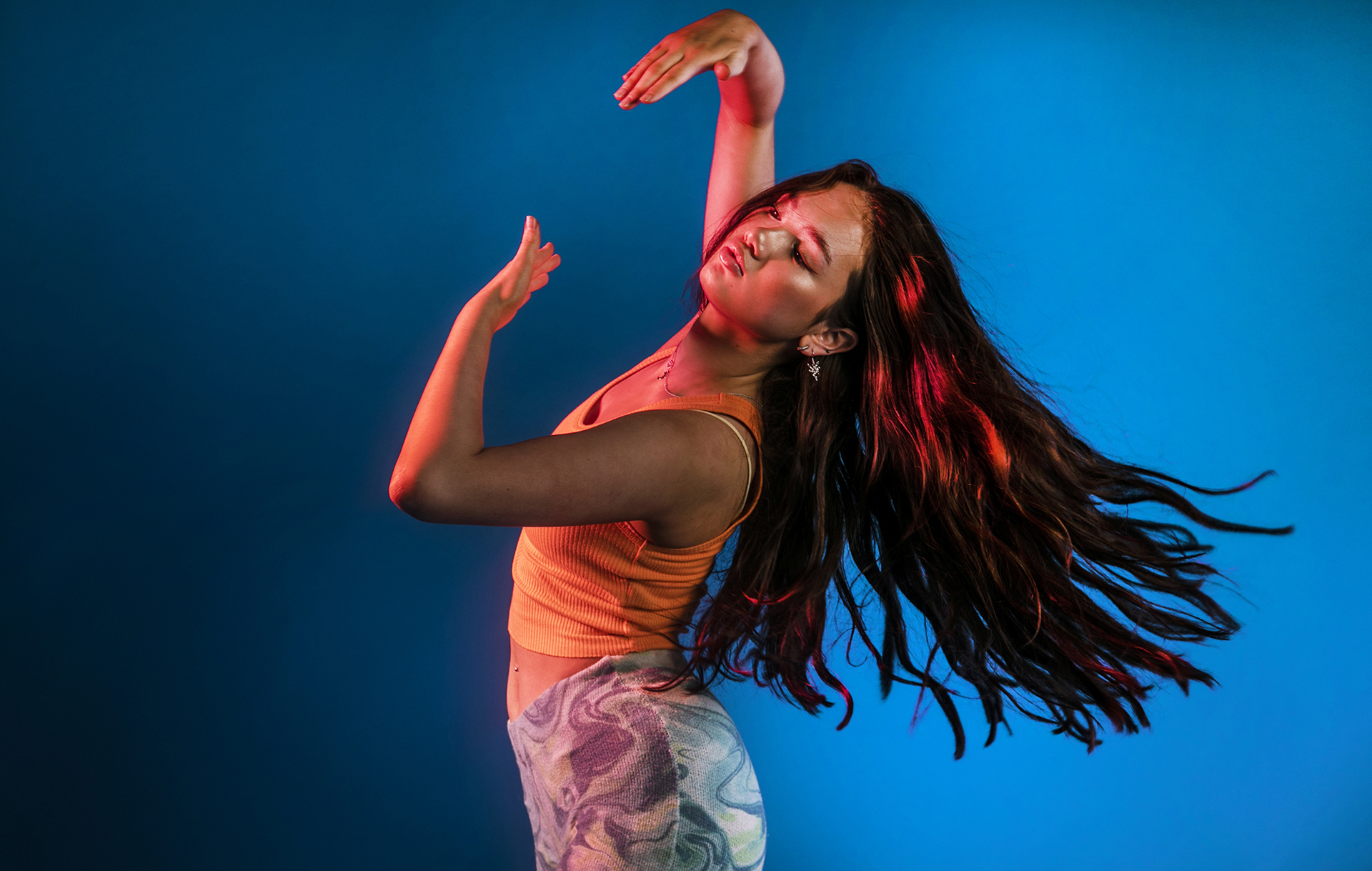 global majority dancer flicking long brown hair back with arms infront of face on blue studio background. Wearing orange crop top and camo jeans.