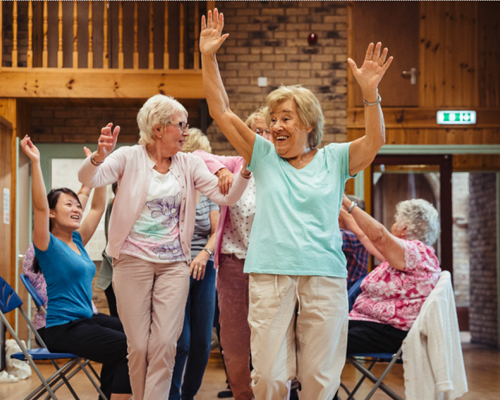 three older white women standing with hands in the air in the centre of the image with two women sitting down with hands in the air either side all taking part in a dance class in a wooden dance studio. 