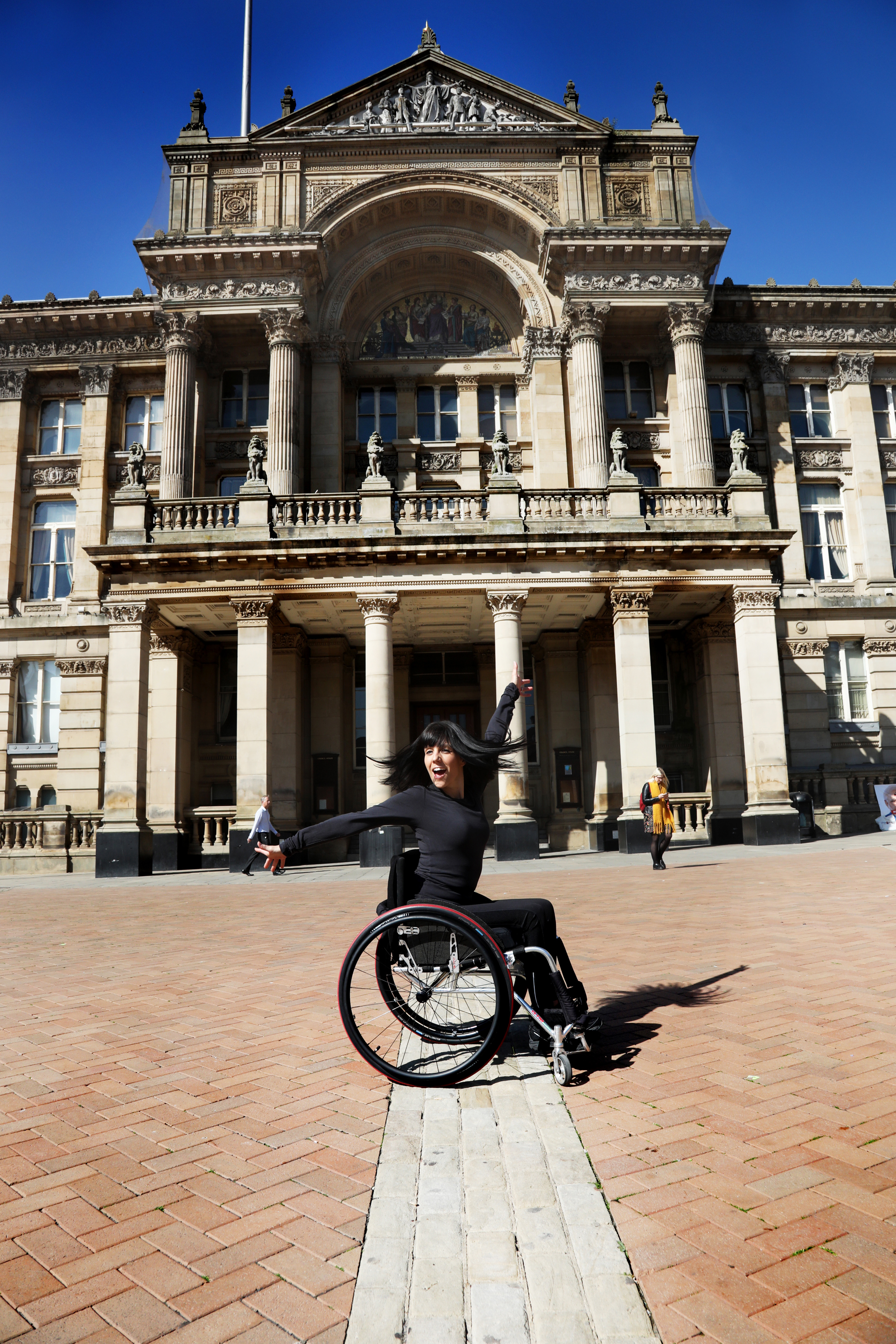 White brunette female dancer wearing all black in a wheel chair flicking her hair with one arms stretched above her head and the other out to the side. Outside a large pillared building. 