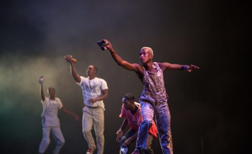four male global majority dancers on dark stage with one arm in the air holding a cloth. 
