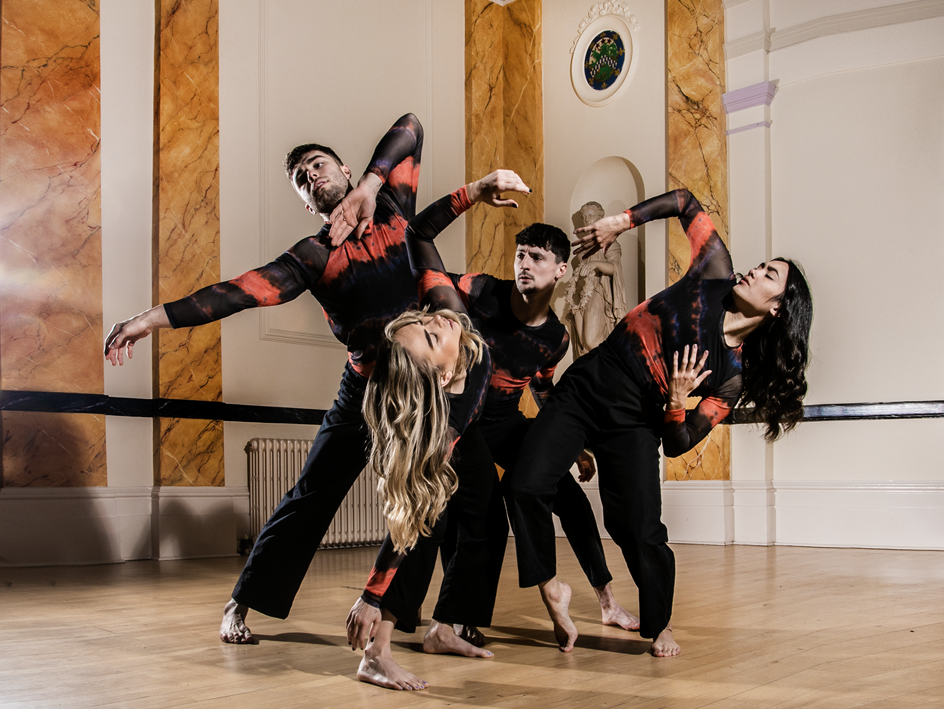 Four dancers intertwined with arms twisted around eachother in a museum building. two white male dancers 2 white female dancers all wearing black and red long sleve tops and trousers.  