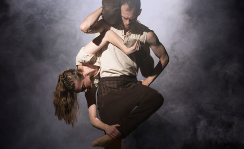 white male dancer standing straight with a white female dancer upside down wrapped around his body, both wearing white tops and black trousers on a smokey dark stage