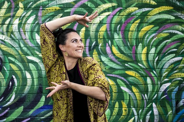 Global majority female dancer with long black hair smiling with arms framing the face. In front of green colourful graffiti wall. Wearing green and purple kaftan. 