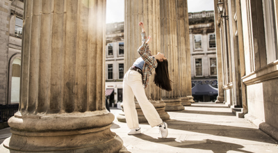 Young female dancer infront of Glasgow coucil building with large pillars. Legs lunched arms above the head. White female dance with long brown hair wearing checked shirt blue top and white trousers 