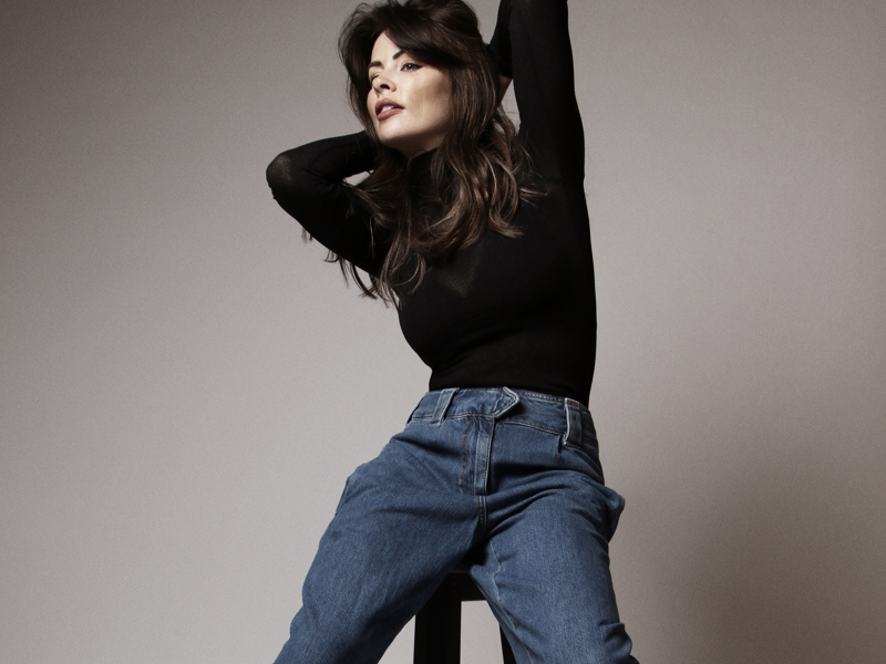 Headshot of Taira Foo. White brown haired female holding elbows in the air with hands behind the head. Wearing black top and blue jeans sitting on high stool