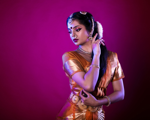 South Asian dancer holding one hand to face and the other towards her elbow on hot pink studio backdrop. Wearing traditional orange dress with gold belt bracelets necklace and head jewlery with gold nose ring.
