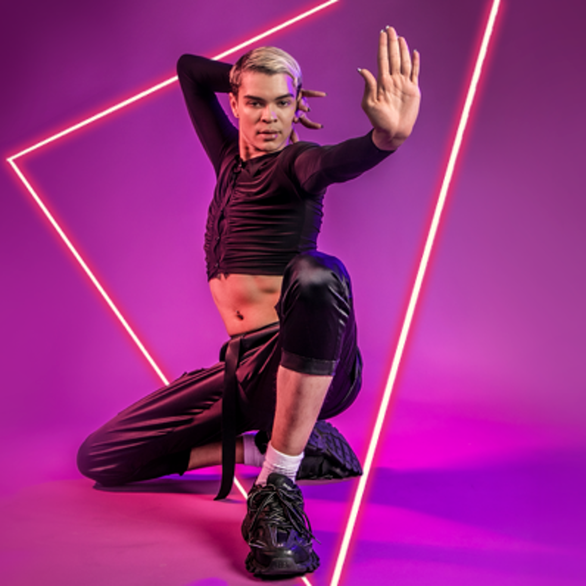 Global majority male LGBTQ+ vogue dancer with slick back blonde hair, kneeling with one hand behind head and the other hand stretched out gesturing stop. On bright purple photo background.