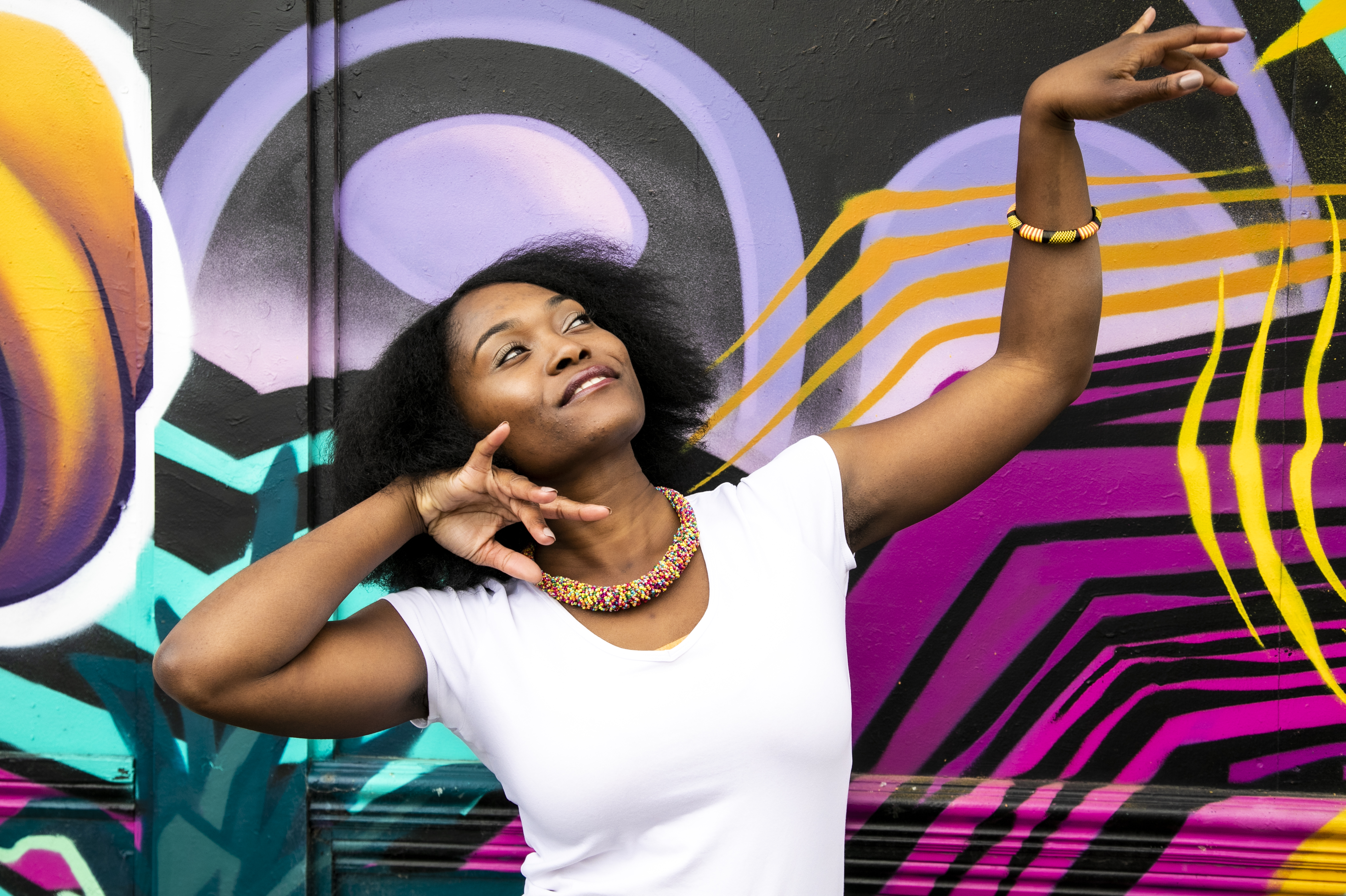 Global majority female dancer with afro, holding one arm out to the top right of the image with eyes looking at a limp wrist and the other hand next to her face. Wearing white top and colourful beaded necklace in fornt of coloured graffiti wall