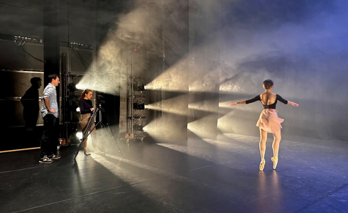 Female dancer on point on stage with stage lights facing her and a two people standing by a camera filming her