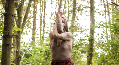 White female crossing tattooed arms over the face in a yoga position. Wearing neutral brown clothes in a woodland area