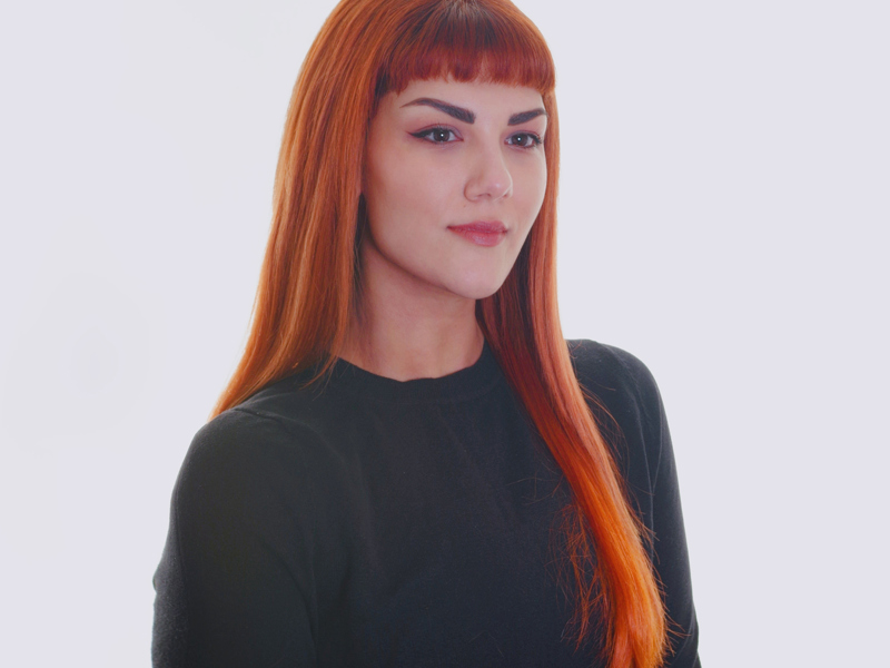 Headshot of Maria Nikolopoulou. Female with long red hair with fringe looking away from the camera. Wearing long sleve black top