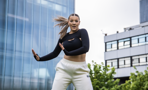 young female dancer with arms to the right side of her infront of Newcastle college building. Wearing black top, white joggers and ponytail swinging to the left