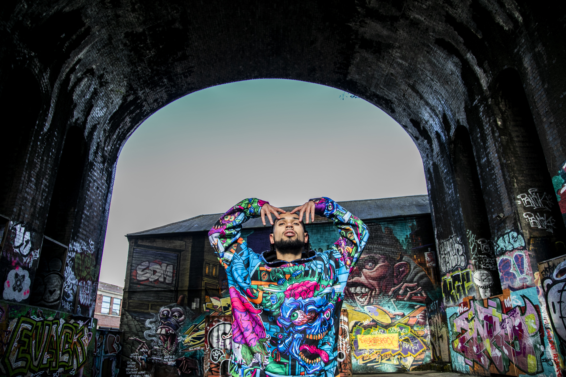 Global majority male dancer with short dreads in colourful blue toned graffiti jumper. with arm in a M shape holding his face and looking up. In front of graffiti building in a large graffiti archway.