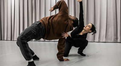 two dancers one male global majority dancer wearing brown hoodie and black joggers healing back in a bridge with one elbow pointed up. one white female wearing full black supporting other dancer with one hand and reaching up with the other