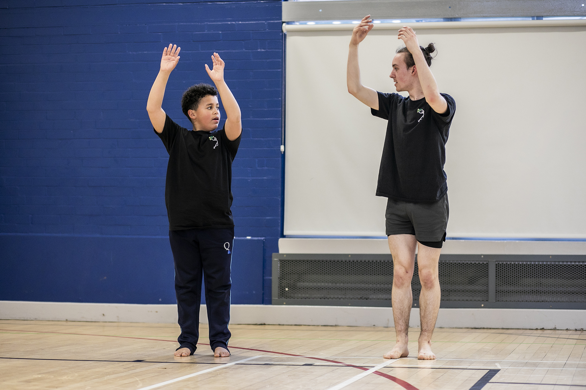 teenage male dance teacher reaching up to the sky with both arms teaching young boy dancer who is mirroring the arms in the air. In sports hall both wearing black PE kit