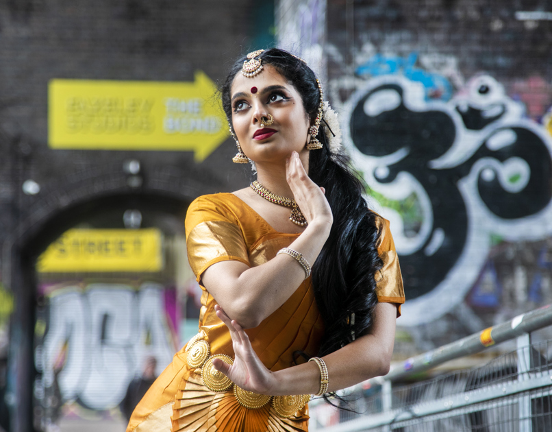 South Asian dancer wearing traditional gold and dress with gold jewellery headpiece. Dancer with one had framing the face and other touching the elbow . In front of graffiti wall