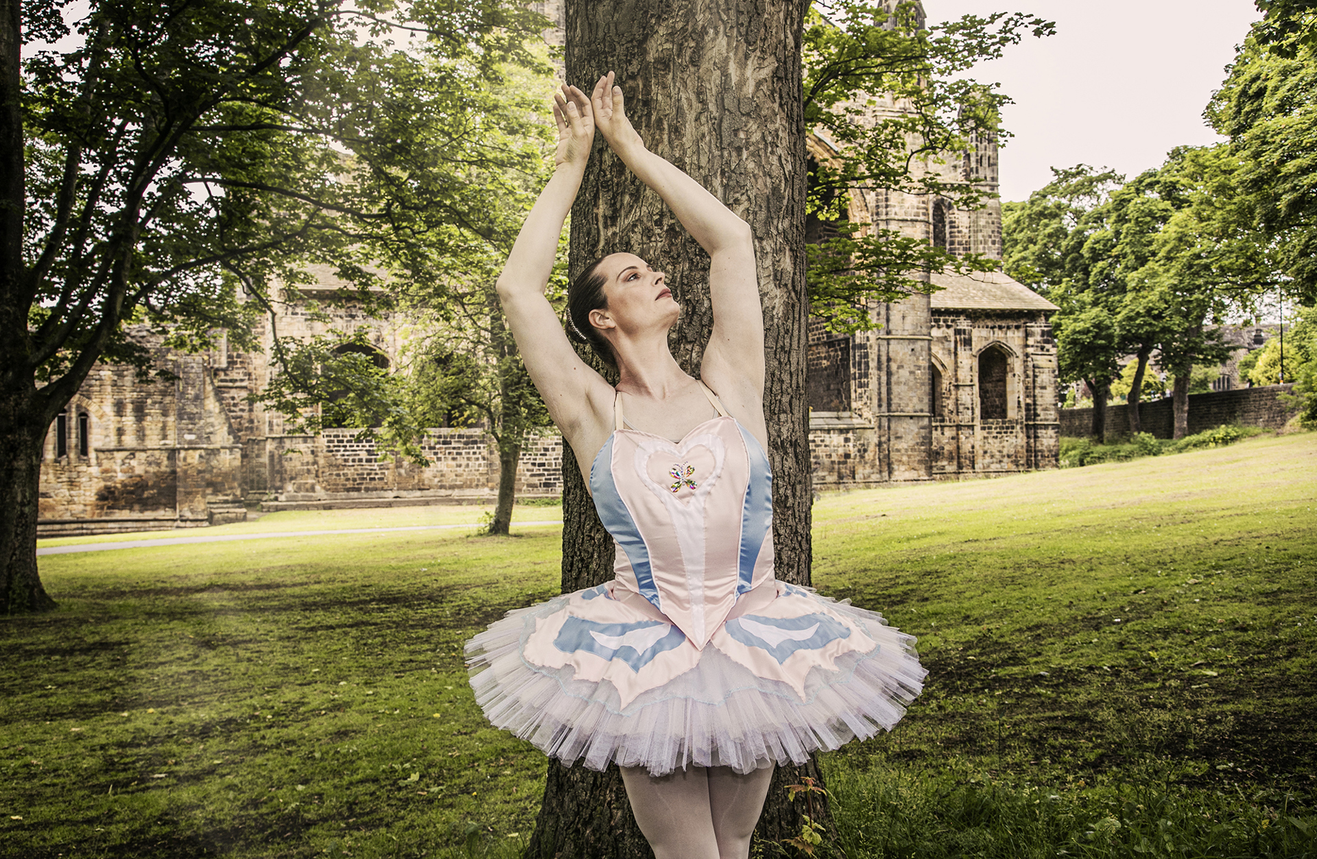 Transgender female ballera wearing Trans flag tutu, arms raised above her head in front of tree and old building in the background