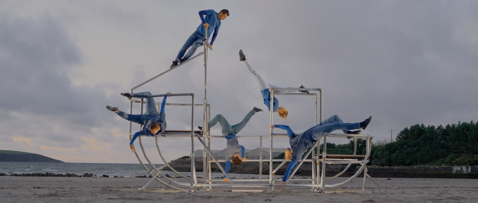 five dancers moving a metal shaped boat scaffolding structure on a beach. All wearing blue and grey outfits five dancers moving a metal shaped boat scaffolding structure on a beach. All wearing blue and grey outfits