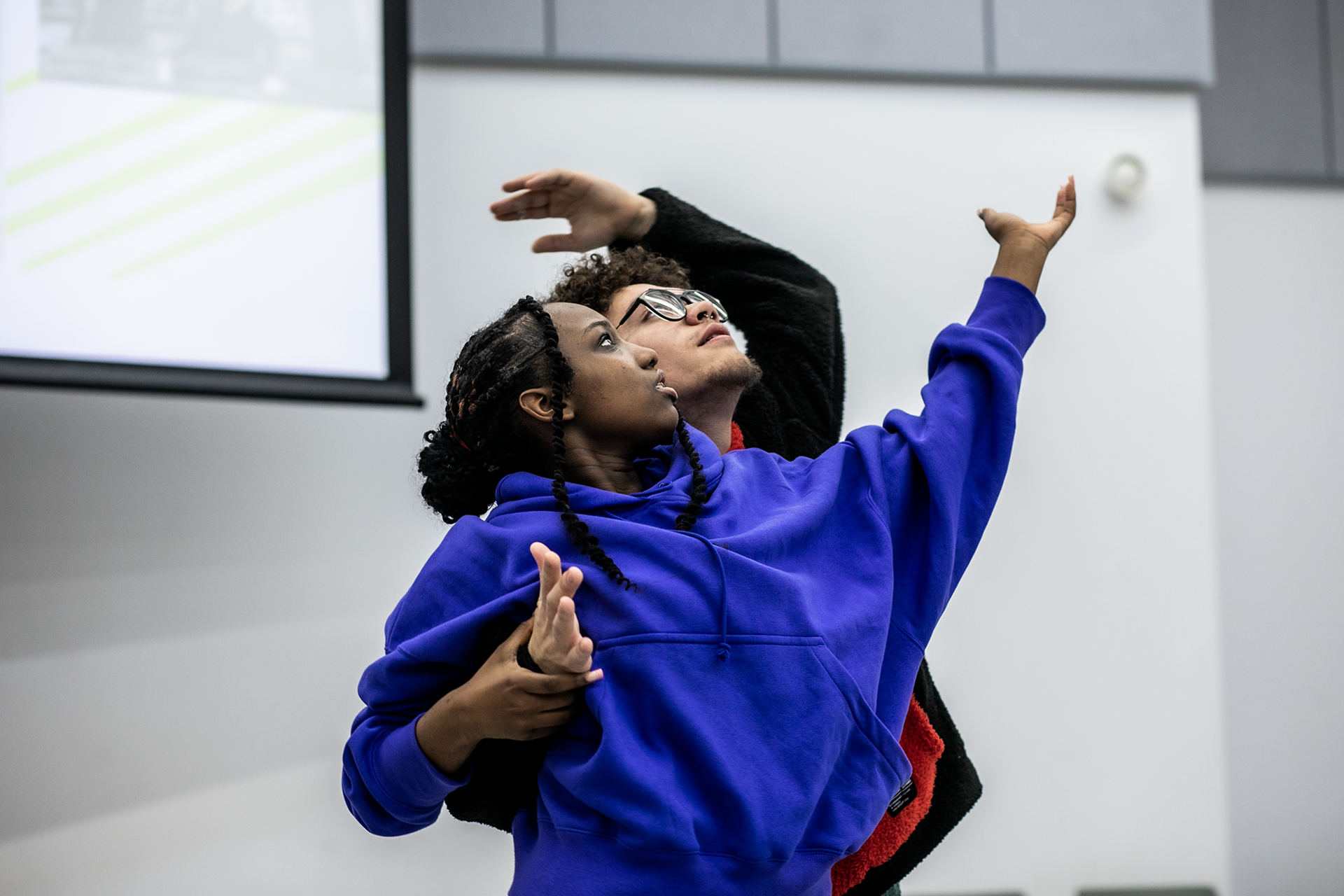 Two dancers, one global majority female with one arm high wearing purple hoodie and a male global majority dancer hugged around female with arm reaching to the top right wearing wholly black jumper. 