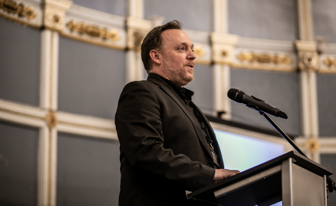 White male presenting in a large historical looking room in front of plinth and microphone. 