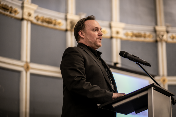 White male presenting in a large historical looking room in front of plinth and microphone. 