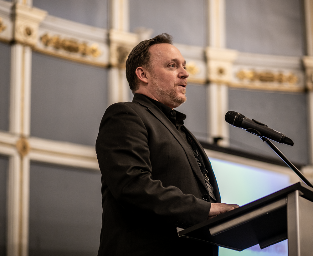 White male presenting in a large historical looking room in front of plinth and microphone.