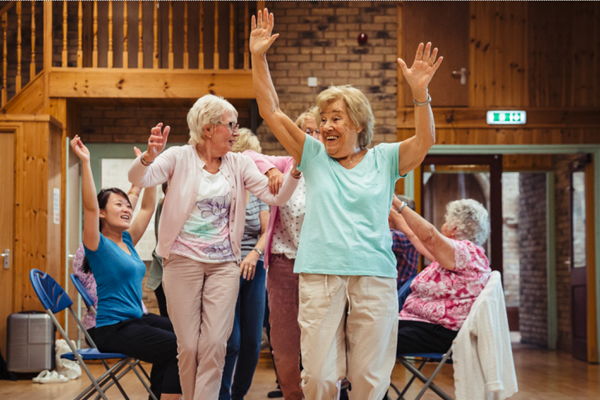 three older white women standing with hands in the air in the centre of the image with two women sitting down with hands in the air either side all taking part in a dance class in a wooden dance studio. 