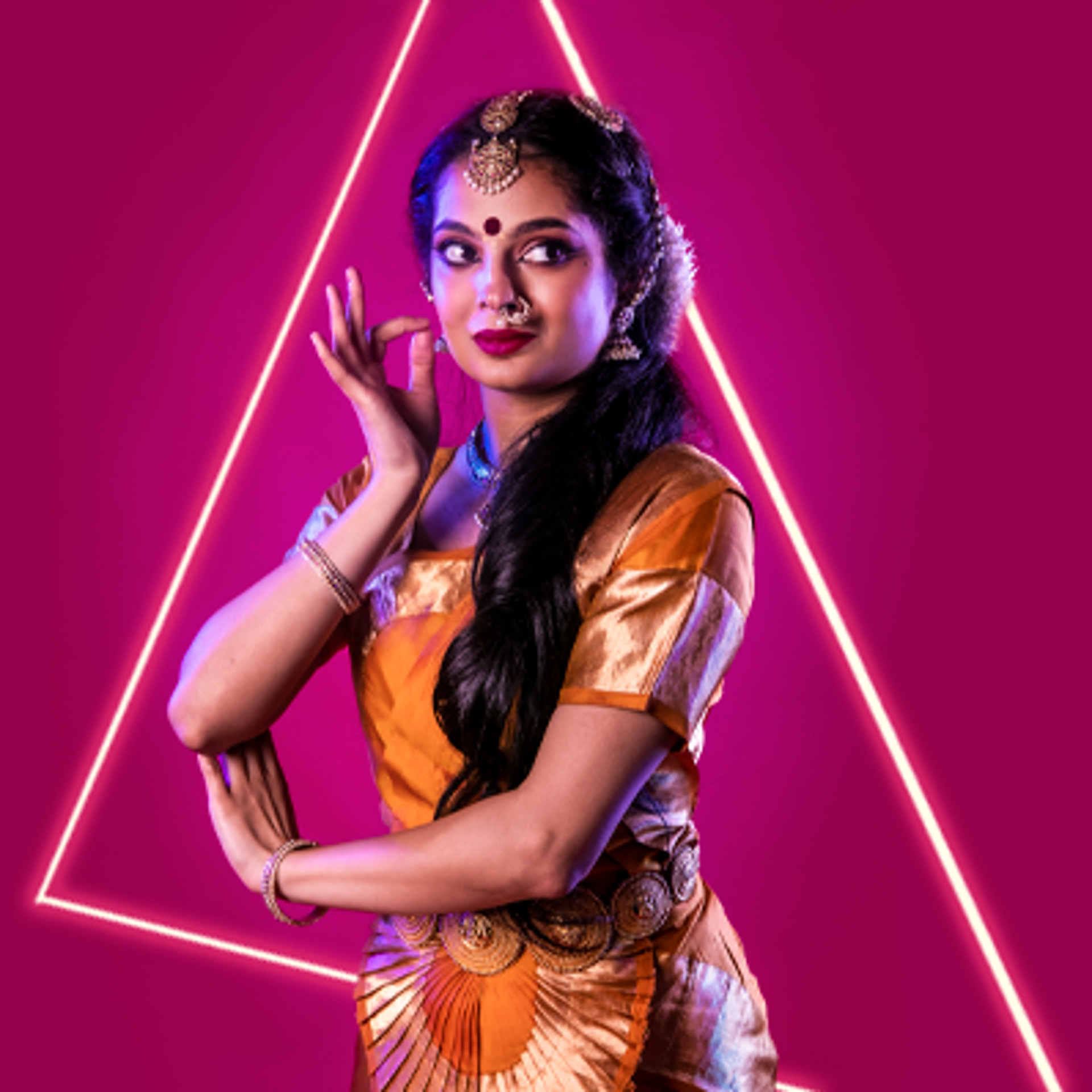 South Asian dancer holding one hand to face and the other towards her elbow on hot pink studio backdrop. Wearing traditional orange dress with gold belt bracelets necklace and head jewellery with gold nose ring.