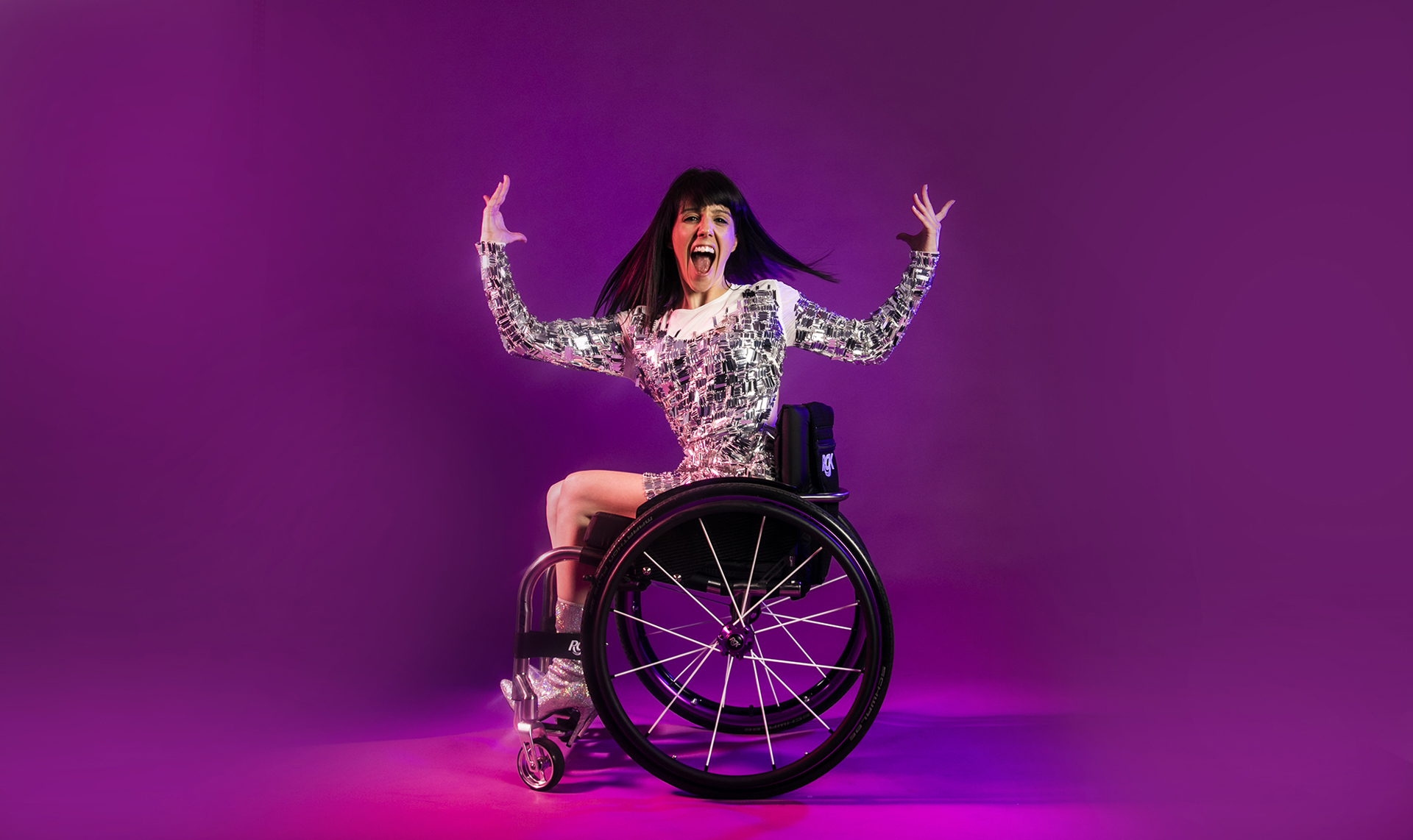 White female wheelchair dancer with black hair. With arms above her head and wheel chair side on. Facial expression wide mouth excited. Wearing silver sparkle dress and silver high heeled shoes in front of hot pink studio background. White female wheelchair dancer with black hair. With arms above her head and wheel chair side on. Facial expression wide mouth excited. Wearing silver sparkle dress and silver high heeled shoes in front of hot pink studio background.