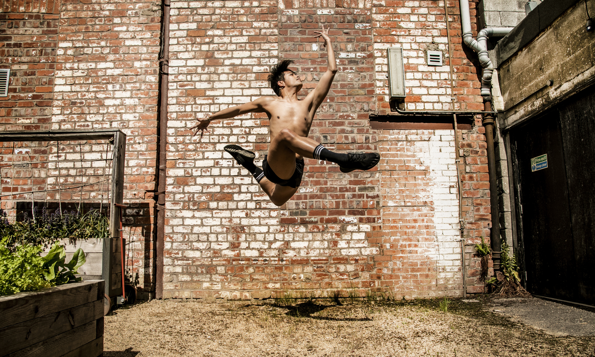 Global majority male dancer jumping high with legs tucked in a running shape, with one arm bent infront of him hand facing the sky and the other arm bent behind him. Wearing no t-shirt and black shorts with black trainers in front of red brick wall 