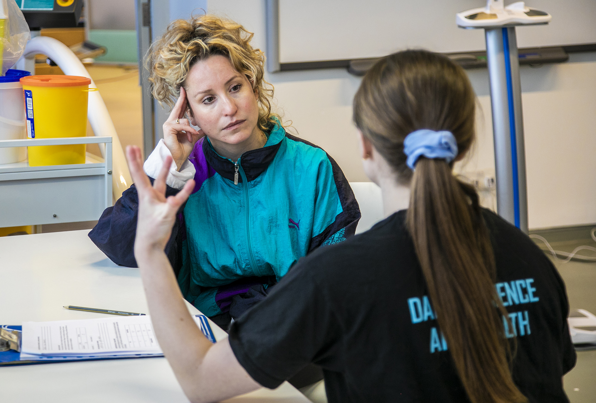 The back of white female with brown tied back hair wearing black sitting at a table gesturing towards white female dancer with curcly blonde hair and blue retro jacket leaning on one hand listening. with clipboard on the table and excersise equipment in the background in white room.