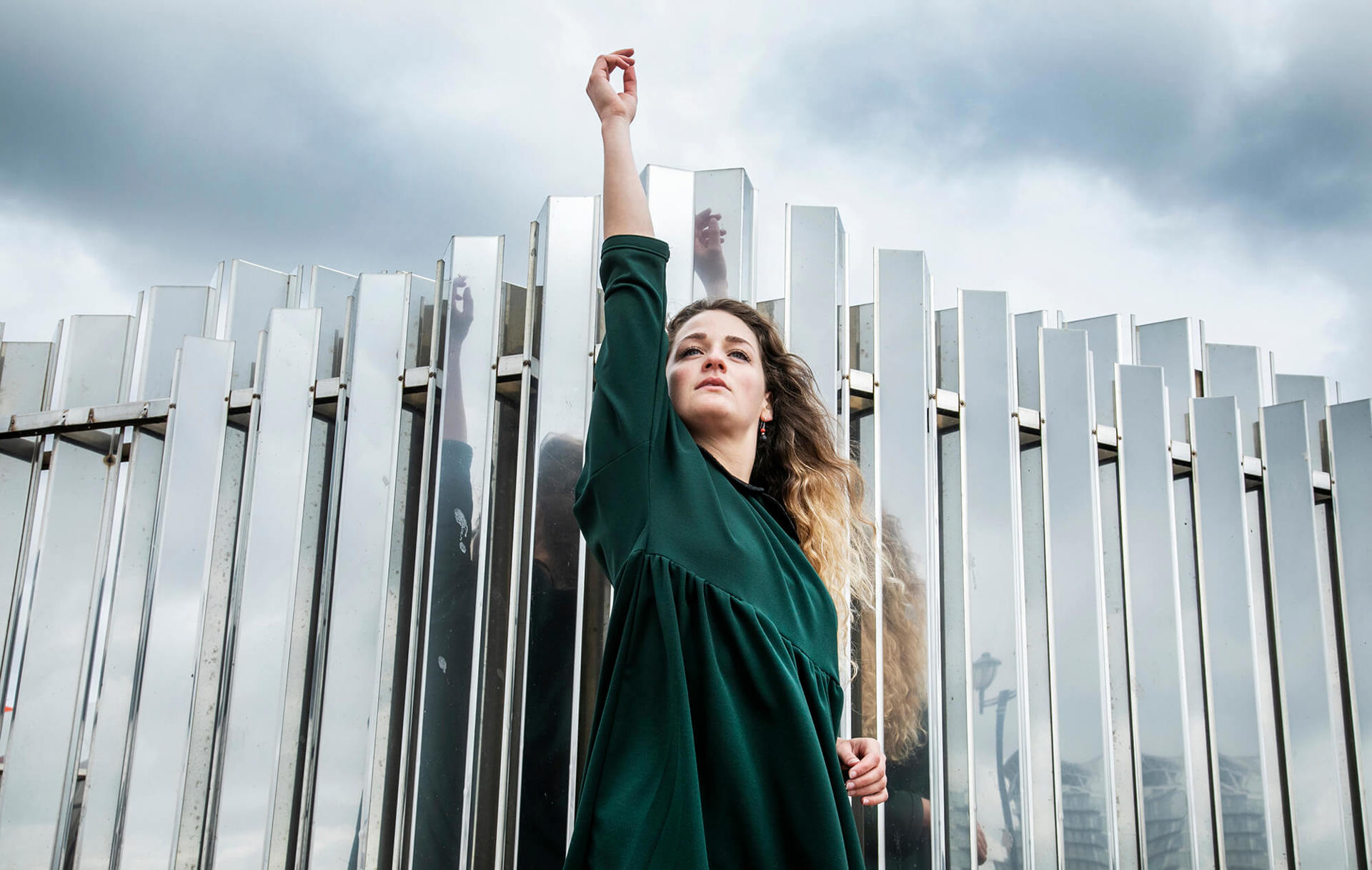dancer standing infront of mirrored wall with stormy sky with one hand in the air. White female with curly long blonde hair and green dress dancer standing infront of mirrored wall with stormy sky with one hand in the air. White female with curly long blonde hair and green dress