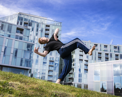 yong white male red haired dancer wearing blue tracksuit bottoms and black t-shirt. leaning back in a running man position on grassey hill in front of Trinity Laban University building 