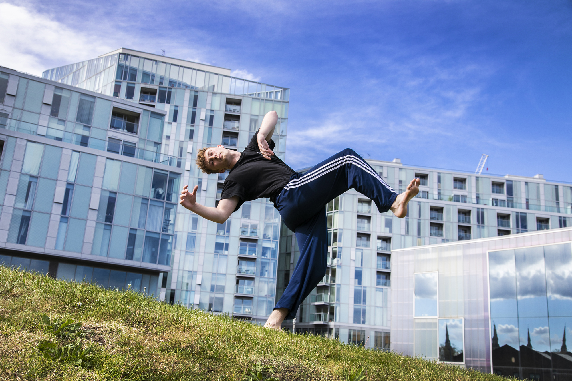 yong white male red haired dancer wearing blue tracksuit bottoms and black t-shirt. leaning back in a running man position on grassey hill in front of Trinity Laban University building 