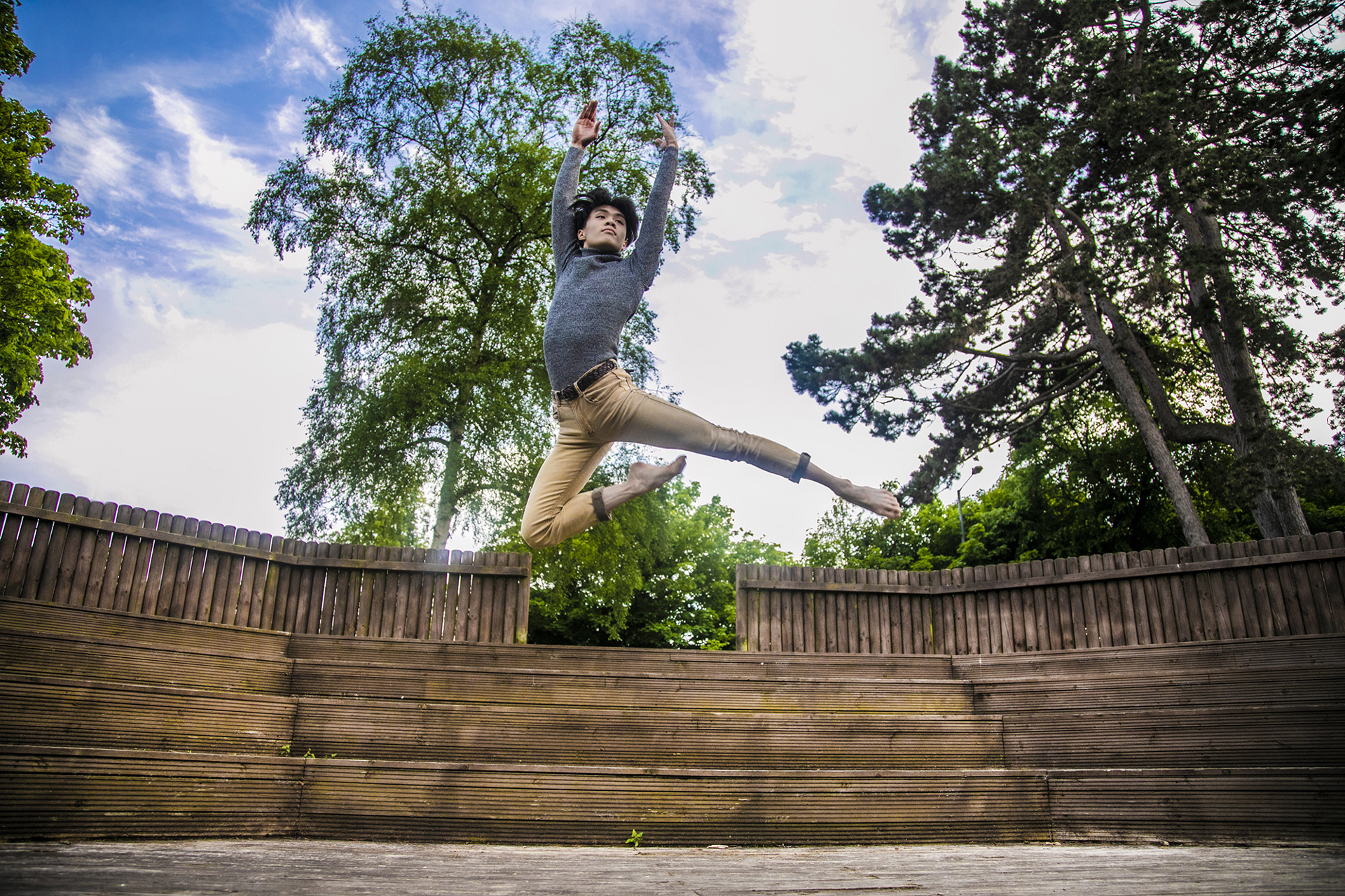 global majority male dancer jumping high with arms above head. Wearing grey long sleve tshirt and beige trousers. In front of soft focus trees and blue sky. global majority male dancer jumping high with arms above head. Wearing grey long sleve tshirt and beige trousers. In front of soft focus trees and blue sky.