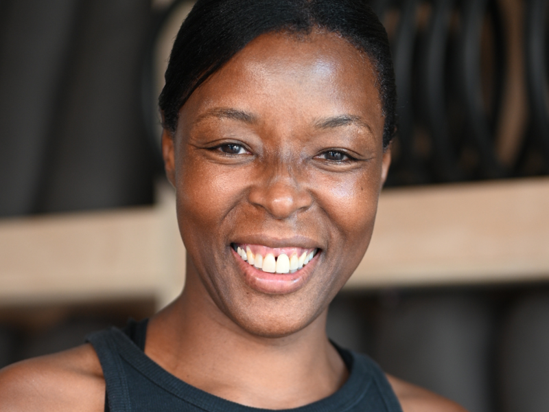 Headshot of Nicolette Wilson Clarke. Global majority female with tied back black hair smiling at camera. Wering black leotard. 