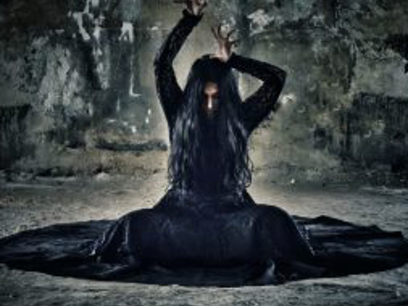 Female wearing all black with hands above head with crossed legs in a gothic room
