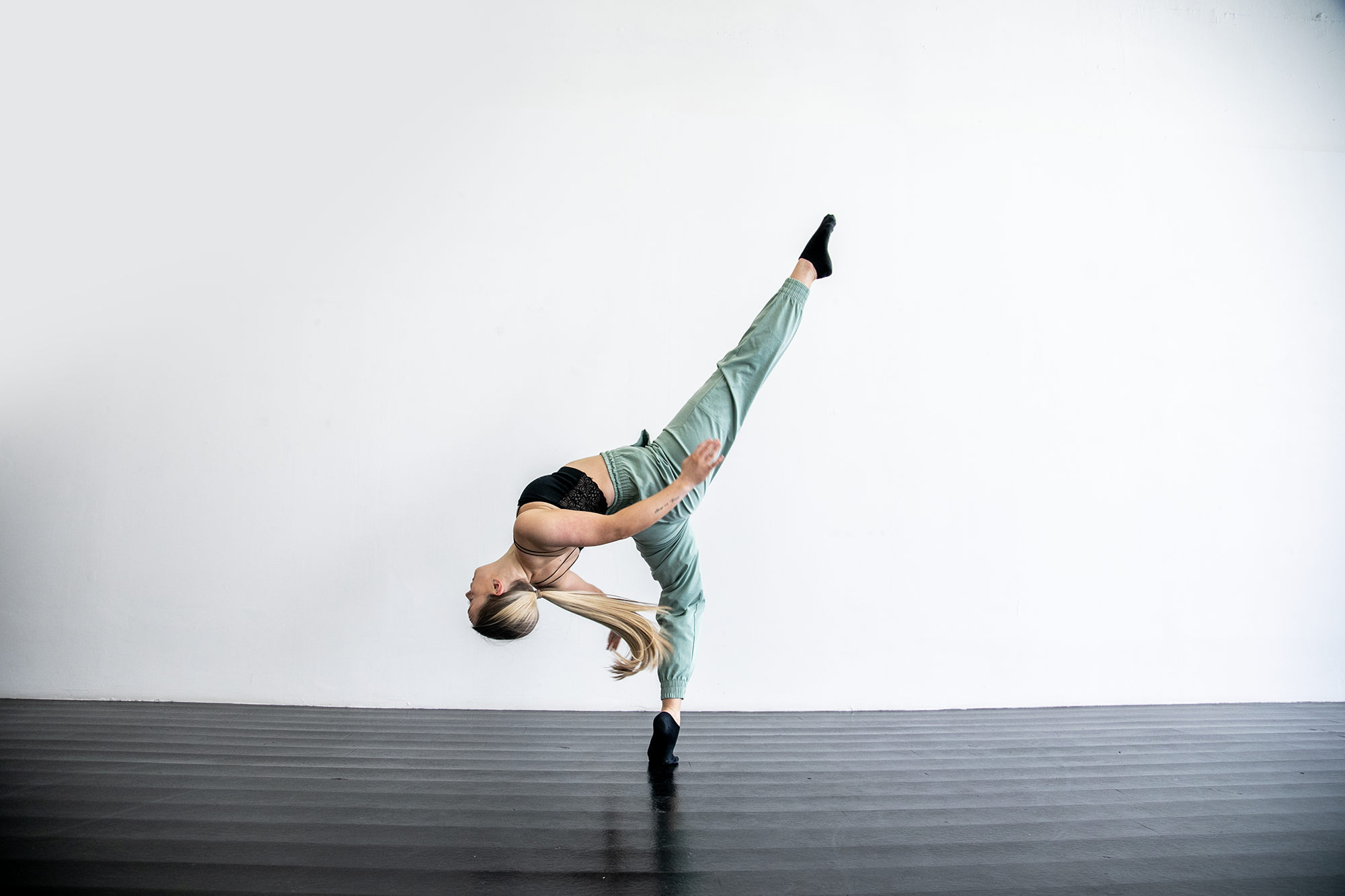 blonde white female in dance studio with black floors. One leg kicked high, other on demi point, leaning back with arms behind. Wearing blak strap top and green trousers and long blonde ponytail 