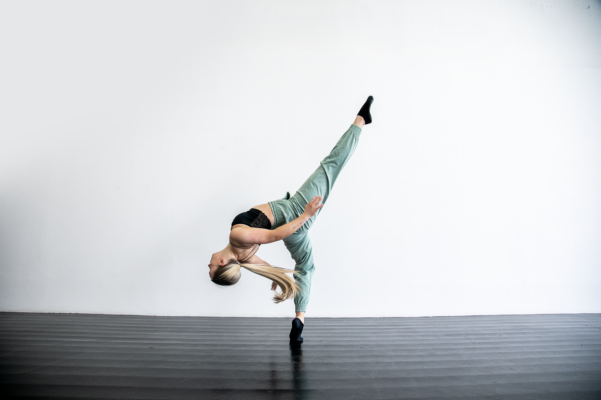 blonde white female in dance studio with black floors. One leg kicked high, other on demi point, leaning back with arms behind. Wearing blak strap top and green trousers and long blonde ponytail 