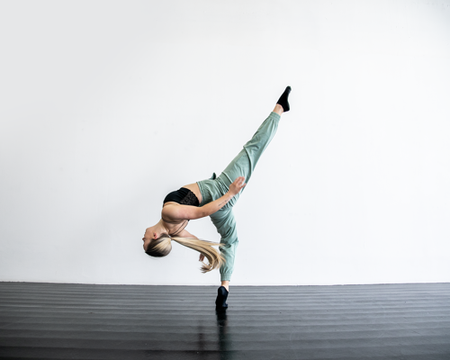 blonde white female in dance studio with black floors. One leg kicked high, other on demi point, leaning back with arms behind. Wearing blak strap top and green trousers and long blonde ponytail 