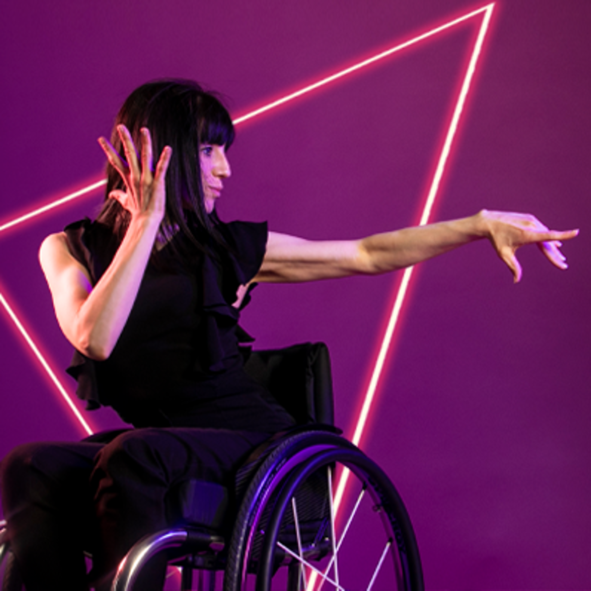 White female wheelchair dancer with black hair. With one hand holding her ear and the other arm pointed to the right hand side of the image, her head and wheel chair side on. Wearing black top and trousers in front of hot pink studio background.