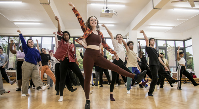 young dancers rehearing in dance studio. All with one one leg up and one arm straight leaning to the left. Mix of male and female dancers with different ethnicities. Wearing comfy casual clothes