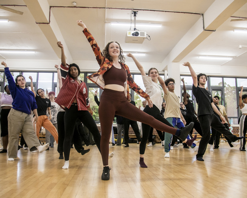 young dancers rehearing in dance studio. All with one one leg up and one arm straight leaning to the left. Mix of male and female dancers with different ethnicities. Wearing comfy casual clothes
