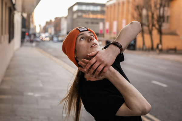 white male dancer with long blonde hair and orage beanie hat and black t shirt. On the street with hand around the face 