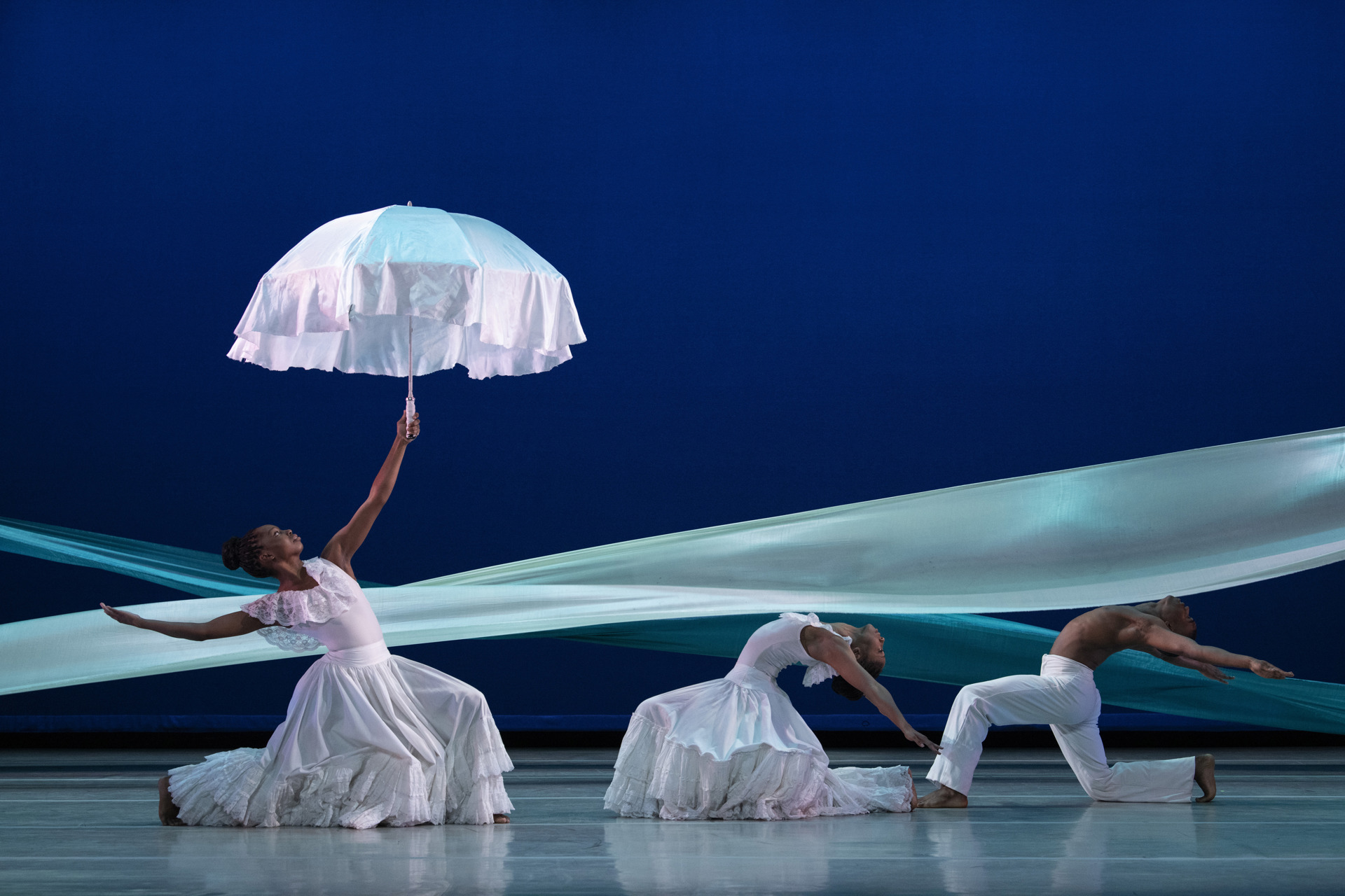three global majority dancers on a blue stage with a blue ribbon graphic behind them. Female to the left lunging and leaning back holding up a white umbrella. Two male and female dancers on the right hand side lunging and arching back. All wearing white.