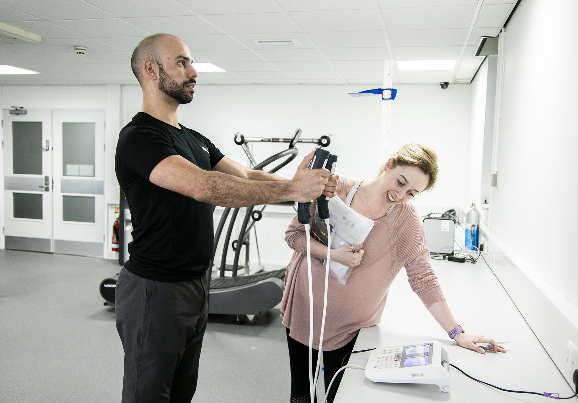 A male dancer is having his body composition measured by standing on a small scale with his arms outstretched in front of him. In either hand he is grasping a body-composition measurement device which are each connected to the scale with white cables. A white female screening assistant is leant over a white desk observing the monitor, and holding some papers. They are in a white and grey dance laboratory, with gym equipment in the background. 