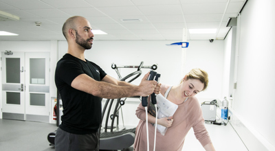 A male dancer is having his body composition measured by standing on a small scale with his arms outstretched in front of him. In either hand he is grasping a body-composition measurement device which are each connected to the scale with white cables. A white female screening assistant is leant over a white desk observing the monitor, and holding some papers. They are in a white and grey dance laboratory, with gym equipment in the background.  A male dancer is having his body composition measured by standing on a small scale with his arms outstretched in front of him. In either hand he is grasping a body-composition measurement device which are each connected to the scale with white cables. A white female screening assistant is leant over a white desk observing the monitor, and holding some papers. They are in a white and grey dance laboratory, with gym equipment in the background.