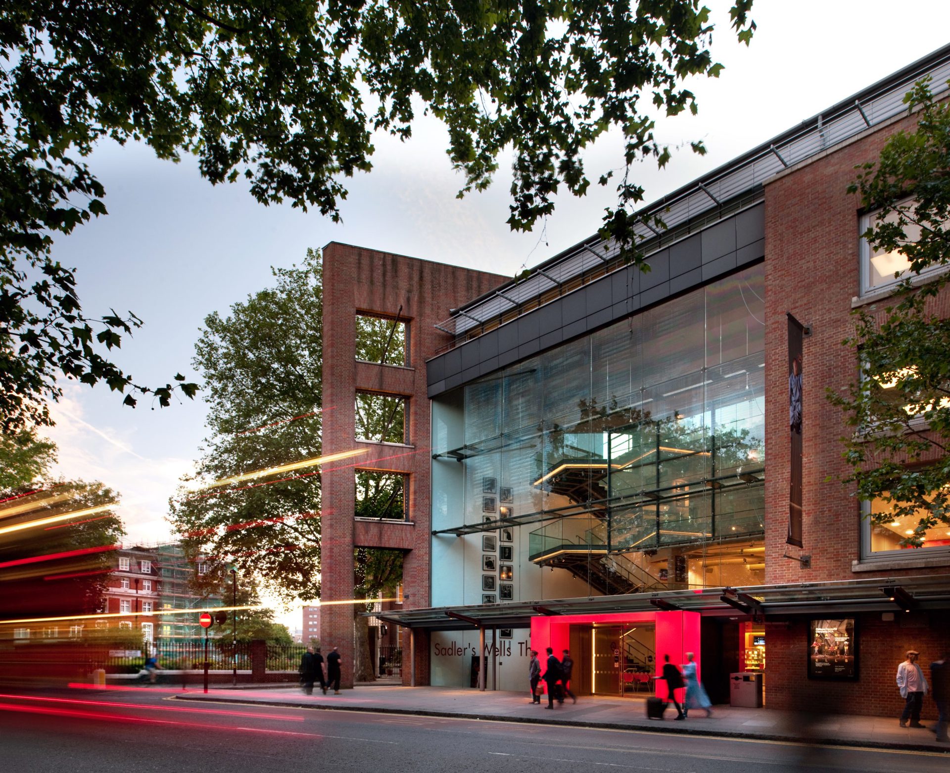 Image of the new Sadlers Wells Theatre. Large brick building with large windows with lit up red door Image of the new Sadlers Wells Theatre. Large brick building with large windows with lit up red door