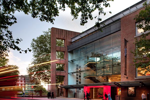 Image of the new Sadlers Wells Theatre. Large brick building with large windows with lit up red door