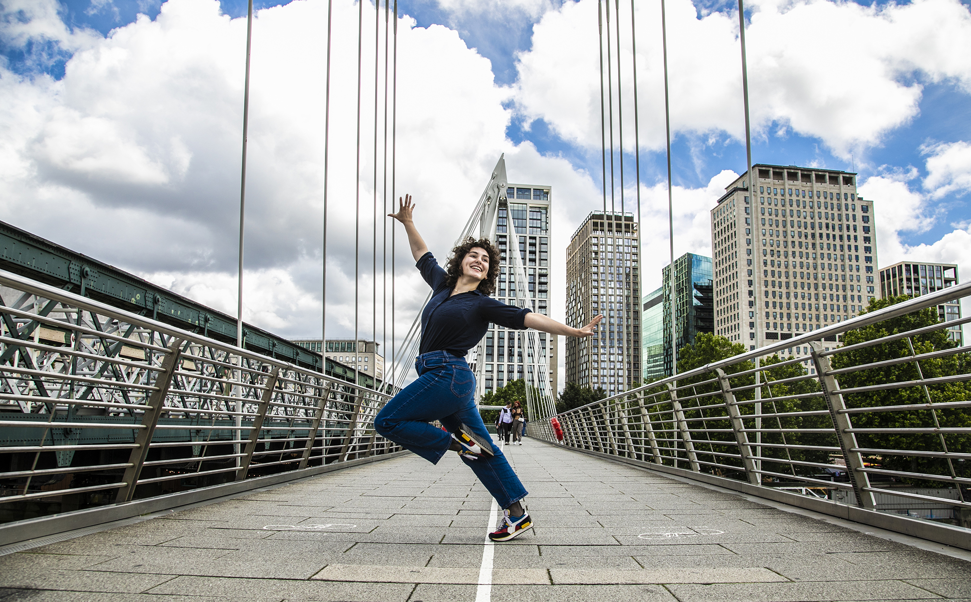 Young dancer with knee up one arm in the air one are to the side dancing on London Bridge. White female with short brown curly hair, wearing black shirt and blue jeans. 